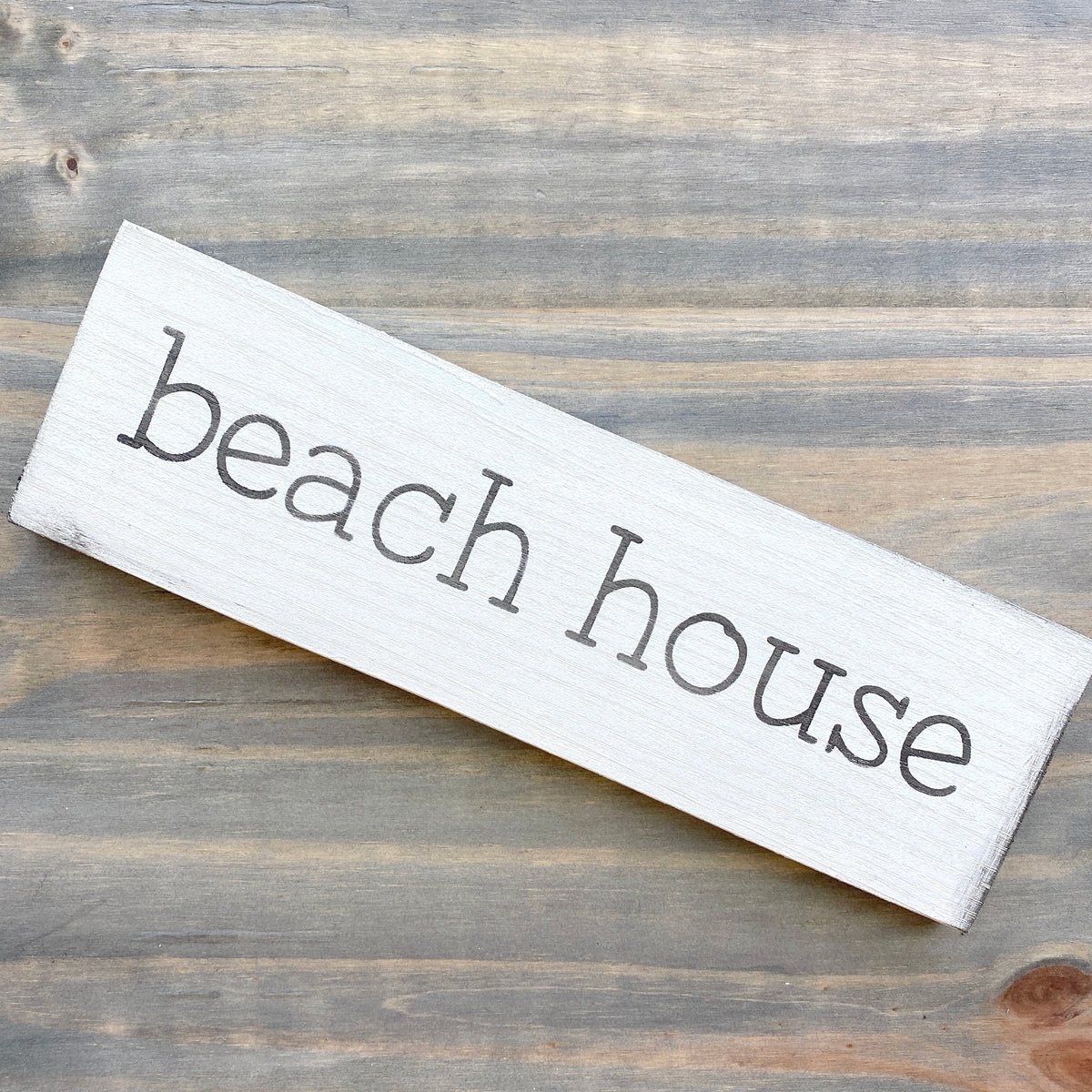 A rectangular white-painted wooden sign with the words 'beach house' in black, placed diagonally on a rustic gray-brown wood plank surface.