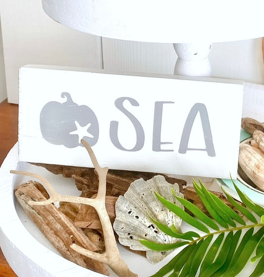 A white rectangular decorative sign with a gray pumpkin, starfish, and the word "SEA," styled on a white tray with driftwood, a shell, and green leaves.