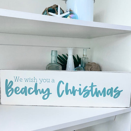 A white rectangular decorative sign on a shelf that reads 'We wish you a Beachy Christmas' in aqua script and serif text. The sign is styled on a white shelf with beach-themed decor, including a white bucket, starfish, green leaves, and glass bottles.