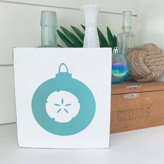 A square white decorative sign featuring a aqua circular ornament design with a white sand dollar in the center, styled on a shelf with glass bottles, green leaves, rope, and a wooden crate in the background.
