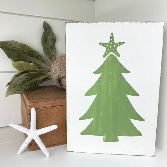 A white rectangular sign featuring a green Christmas tree silhouette with a green starfishon top, styled on a white surface next to a small wooden box, green leaves, and a white starfish.