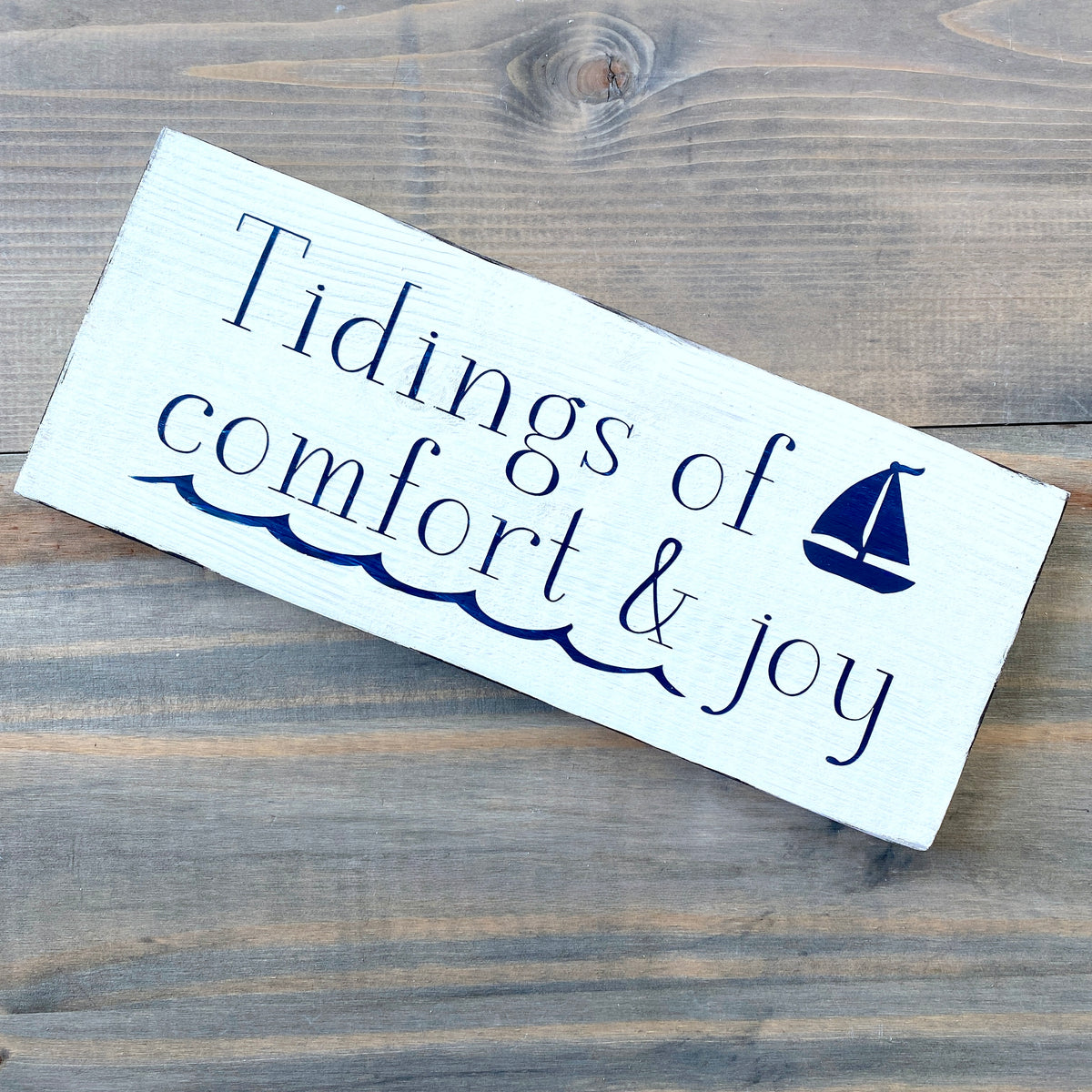 A wooden sign with a white background, featuring the text 'Tidings of comfort & joy' and a small graphic of a sailboat. The sign has a sawtooth hanger and is placed on a wooden surface.