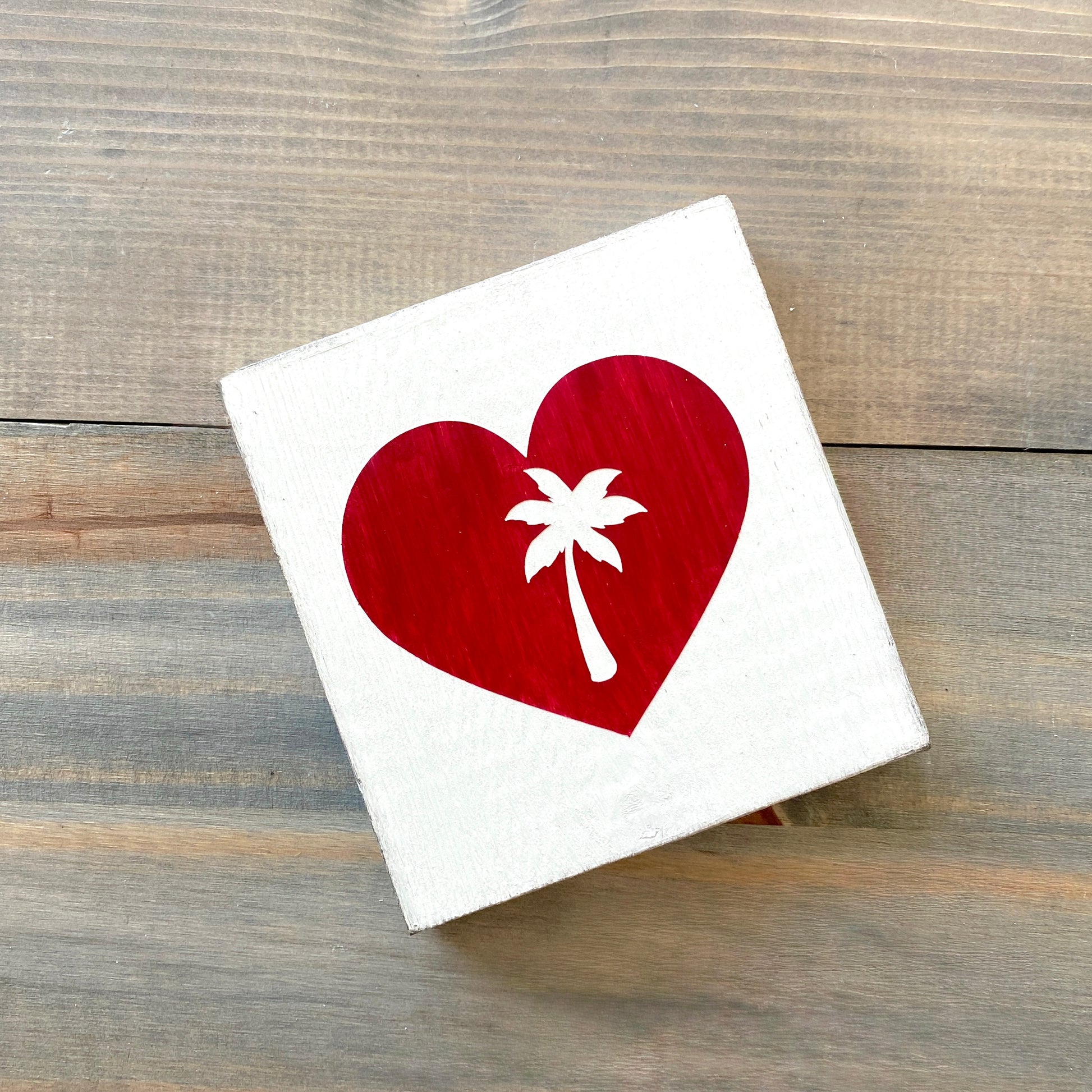 A square white wood sign featuring a red heart with a white palm tree silhouette, displayed on a rustic gray-brown wooden surface.