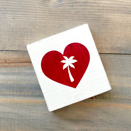A square white wood sign featuring a red heart with a white palm tree silhouette, displayed on a rustic gray-brown wooden surface.