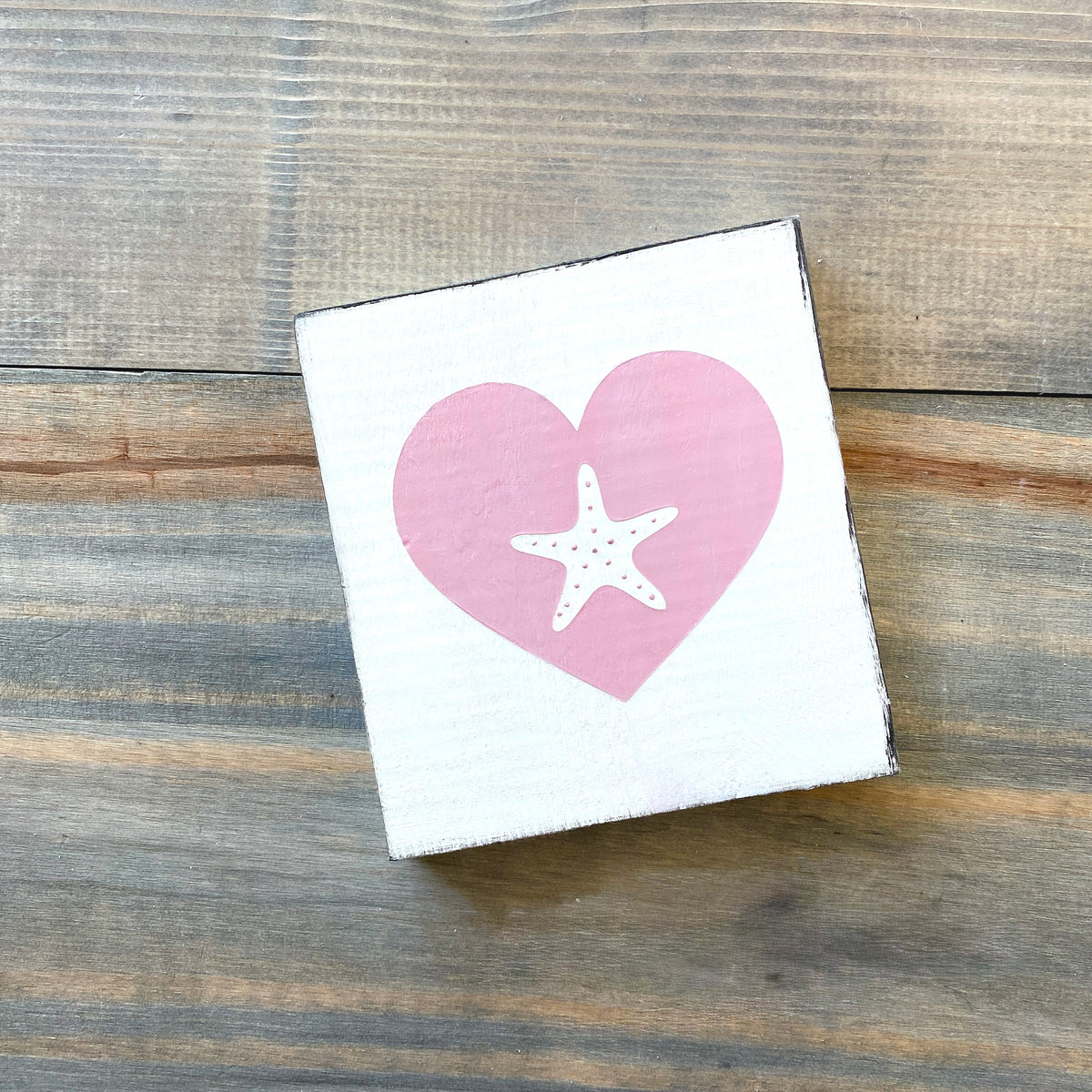A square white sign with a pink heart and white starfish design, photographed on a rustic wood plank surface.