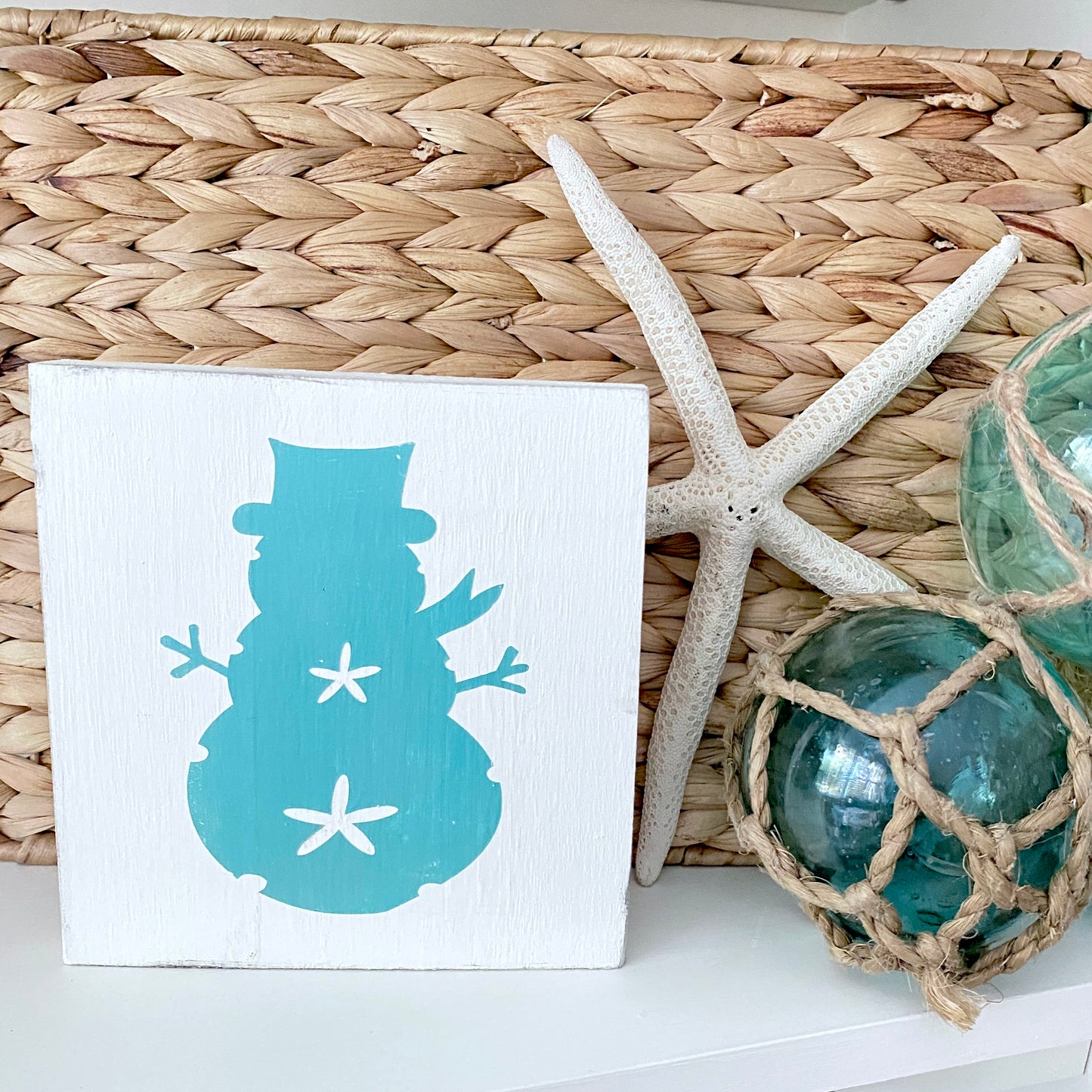 A square white decorative sign featuring a  aqua snowman silhouette with white star accents, styled on a white surface next to a woven basket, a white starfish, and green glass orbs.