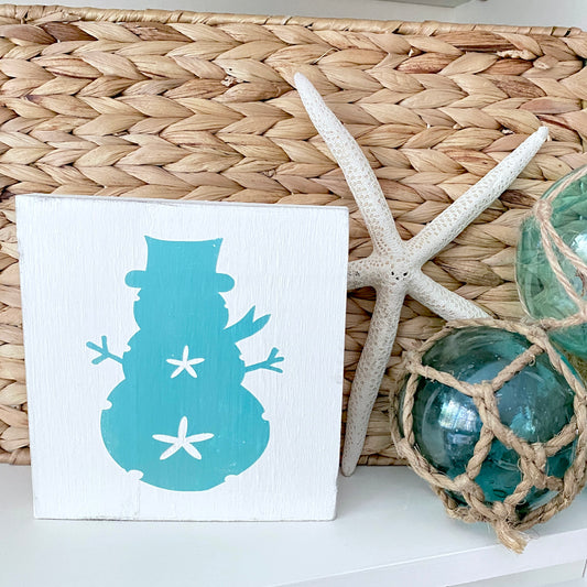 A square white decorative sign featuring a  aqua snowman silhouette with white star accents, styled on a white surface next to a woven basket, a white starfish, and green glass orbs.