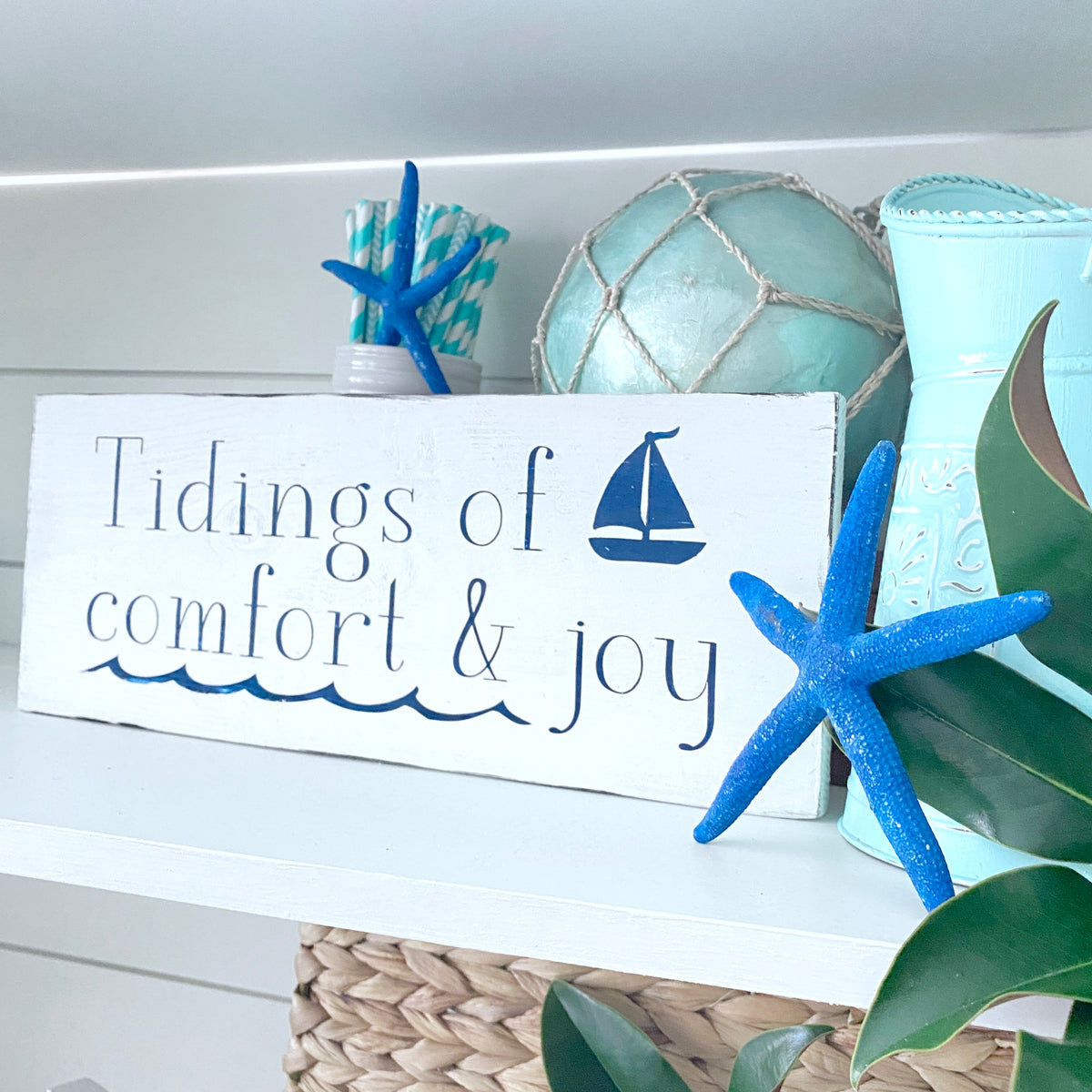 A white rustic wooden sign with the phrase 'Tidings of comfort & joy' in navy blue lettering and a small sailboat graphic, styled on a shelf with blue starfish, a mint fishing float and magnolia leaves.