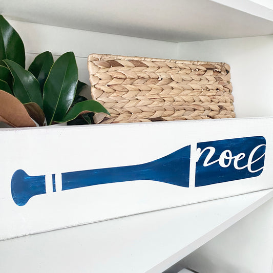 A white wooden shelf with a painted blue oar silhouette and the word 'noel' in white script, styled with a woven basket and green leaves in the background.