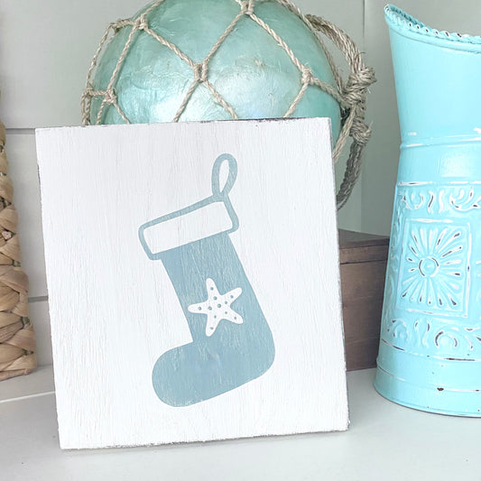 A square white wooden sign featuring a painted sage green Christmas stocking with a white starfish detail, styled on a shelf next to a blue decorative container.