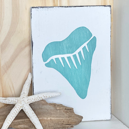 A rectangular wooden sign with a distressed white background and a large aqua shark tooth, styled with a white starfish and driftwood against a light wood wall.