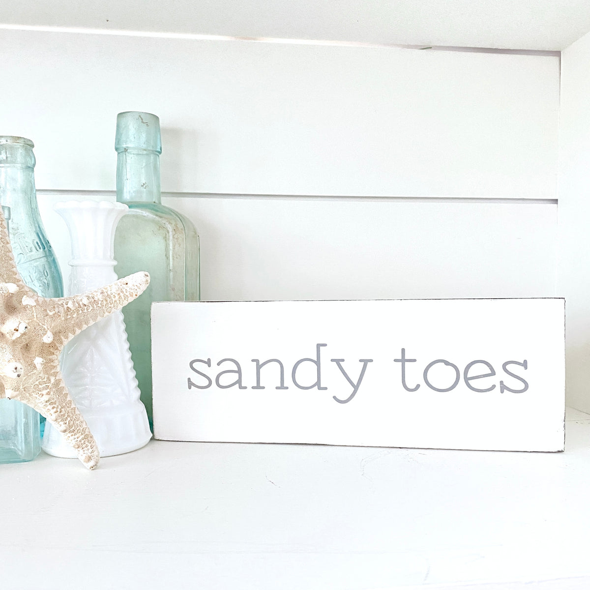 A white rectangular wooden sign with the words 'sandy toes' in gray lowercase font displayed on a white shelf in a styled home decor setting with a wooden box, starfish, and decorative bottles.
