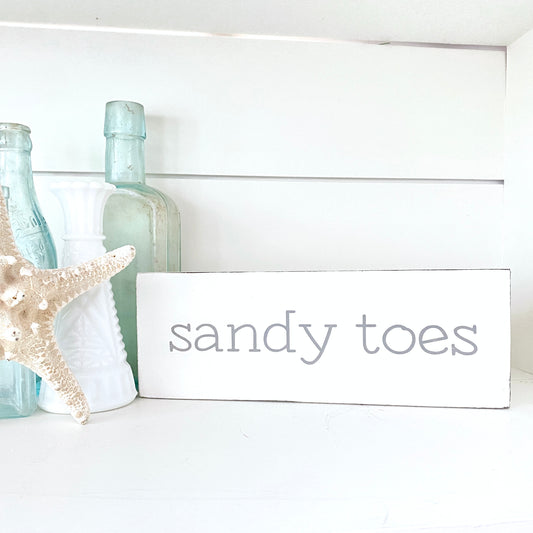A white rectangular wooden sign with the words 'sandy toes' in gray lowercase font displayed on a white shelf in a styled home decor setting with a wooden box, starfish, and decorative bottles.