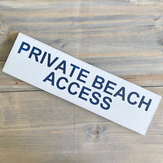 A rectangular white wooden sign with the words 'PRIVATE BEACH ACCESS' printed in dark navy blue, placed diagonally on a rustic wooden surface.