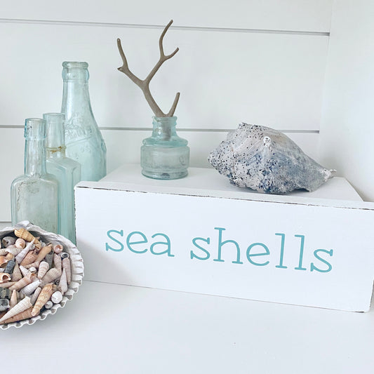 A white decorative sign with the words 'sea shells' in aquae lettering, styled with a gray seashell, small glass bottles, driftwood, and a bowl of assorted seashells on a white surface against a white shiplap wall.