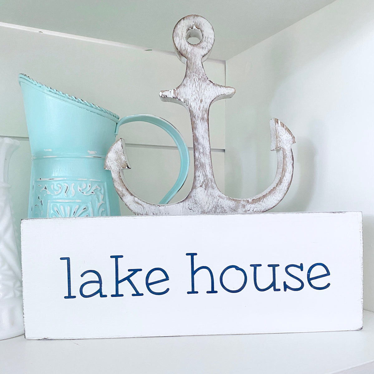 A white rectangular wooden sign with the words 'lake house' in navy, styled on a shelf with a distressed white anchor and a light blue pitcher behind it.