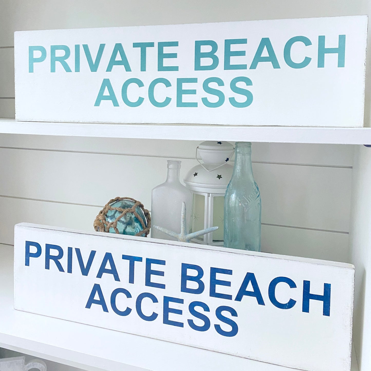 Two white wooden signs with 'PRIVATE BEACH ACCESS' text, in aqua and navy, displayed on a white shelf with decorative coastal decor