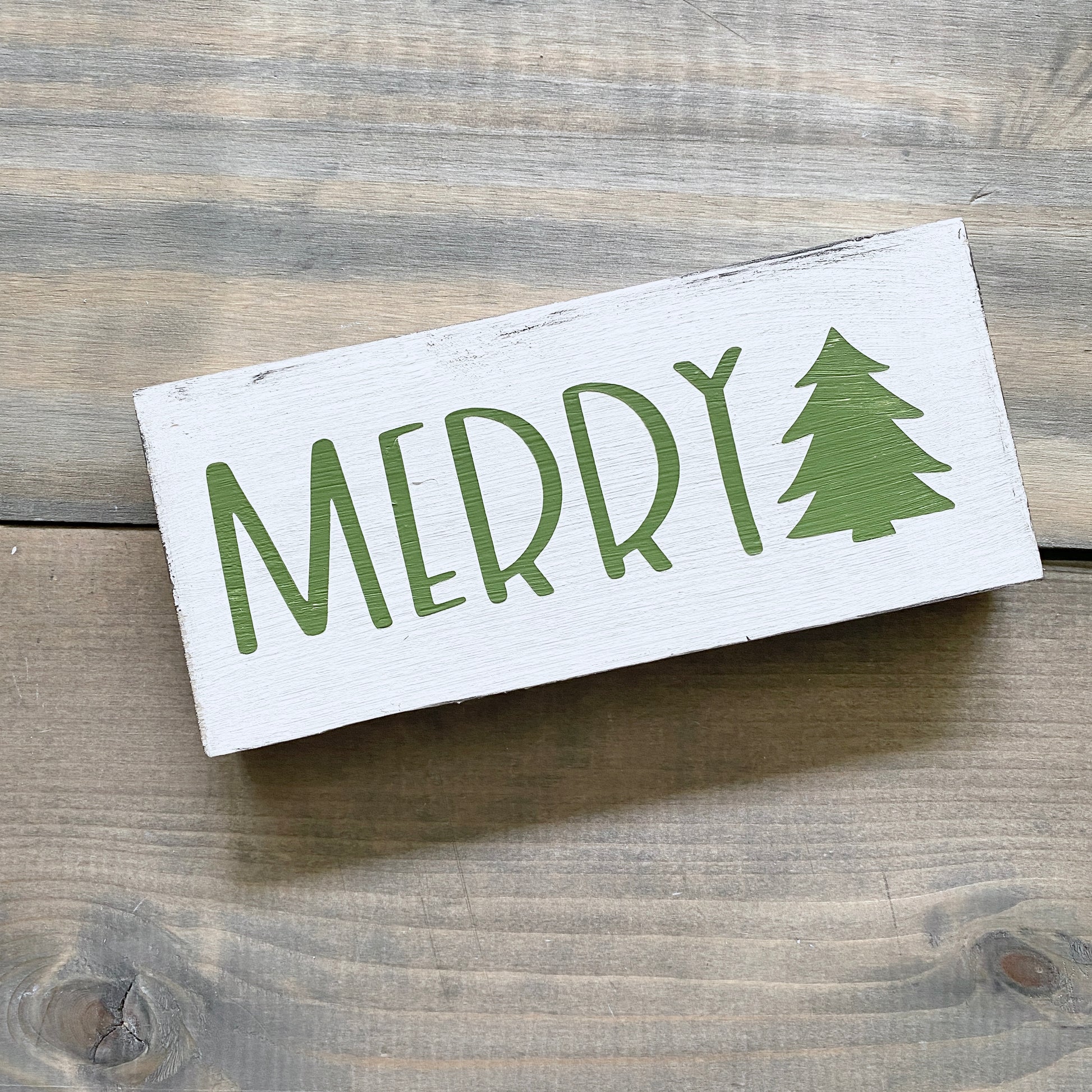 A rectangular white wooden sign with 'MERRY' in green lettering and a green Christmas tree icon, photographed on a rustic wood plank surface.