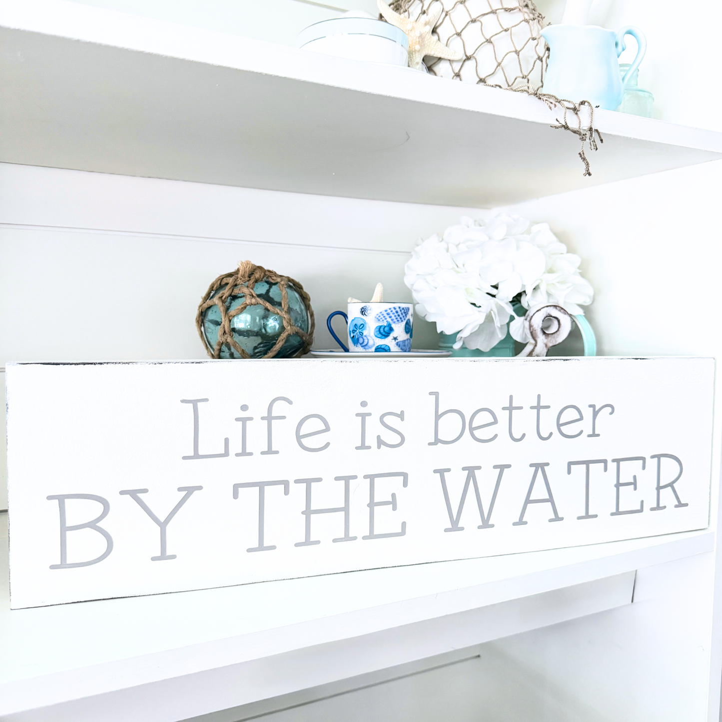 A white rectangular decorative sign with the phrase 'Life is better BY THE WATER' in gray lettering, displayed on a white shelf alongside decorative items including a blue patterned mug, white flowers, and a blue glass orb.