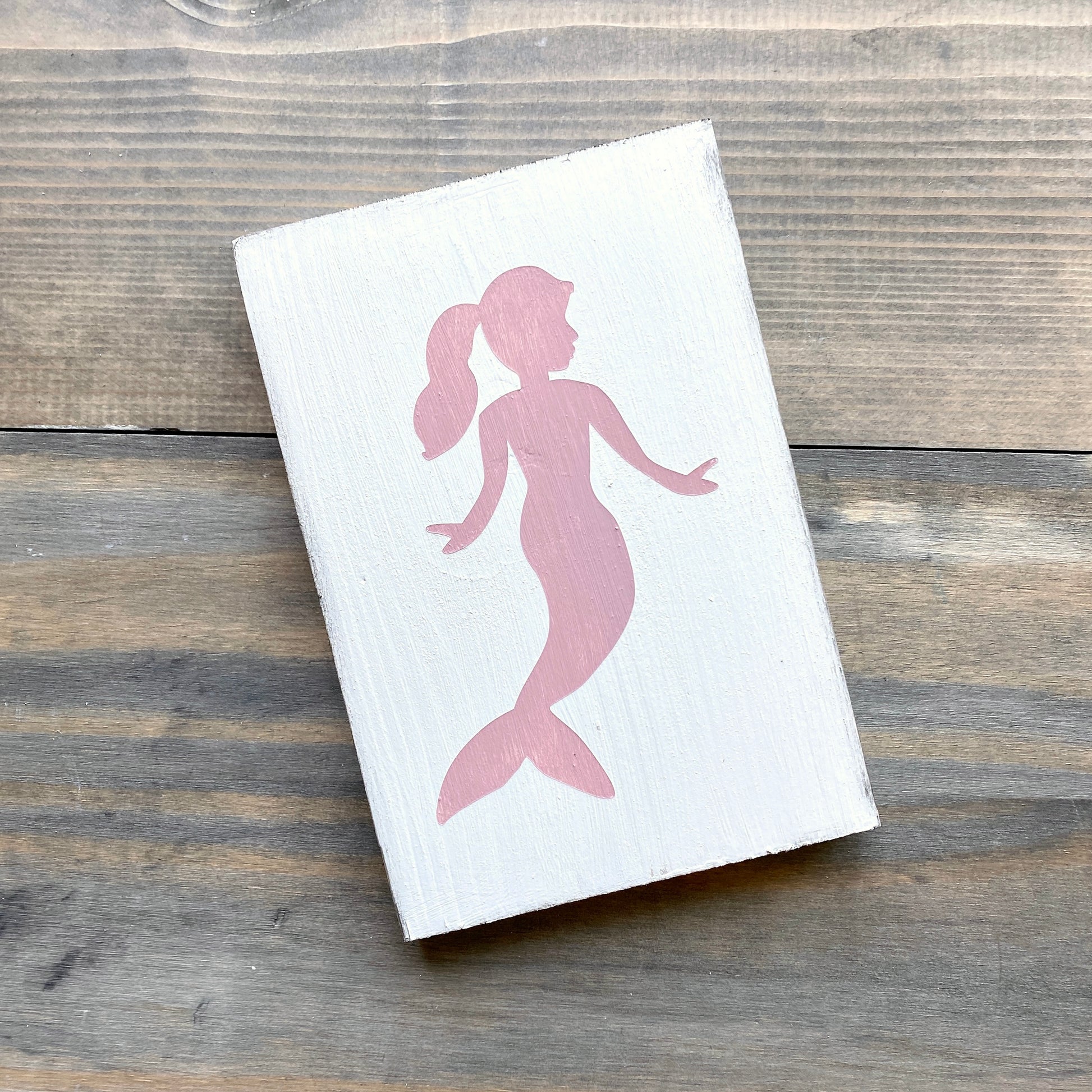 A rectangular white wooden block featuring a pink silhouette of a mermaid, photographed at an angle on a rustic gray-brown wooden surface.