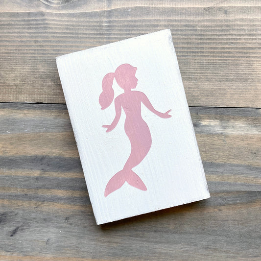 A rectangular white wooden block featuring a pink silhouette of a mermaid, photographed at an angle on a rustic gray-brown wooden surface.