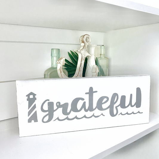 Decorative sign with 'grateful' on a shelf with plants and bottles.