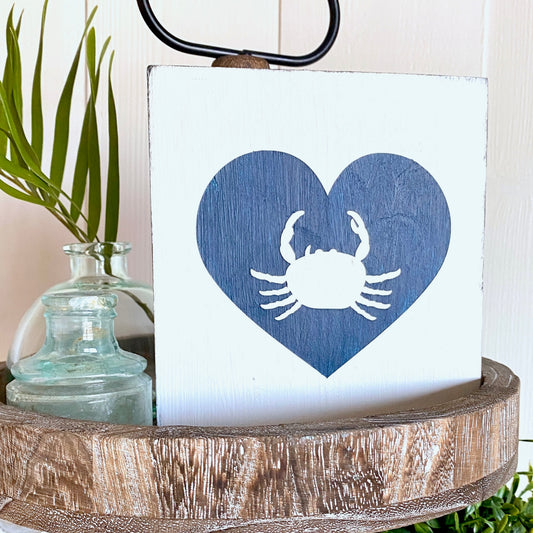Decorative sign with blue heart and white crab design on a wooden surface.