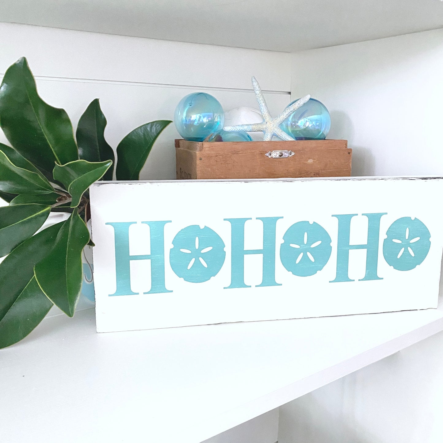 Decorative sign with 'HO HO HO' text on a shelf with plants and decorative items.