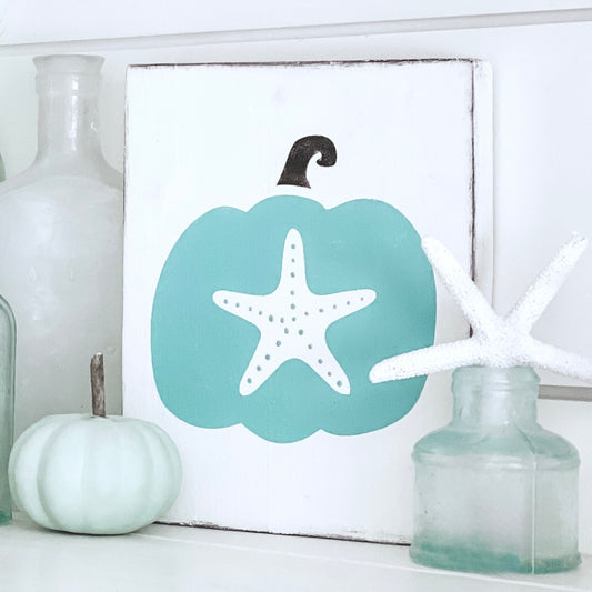 A square white decorative sign featuring an aqua pumpkin silhouette with a white starfish in the center and a small brownstem, styled on a shelf with glass bottles and a small pale green pumpkin.