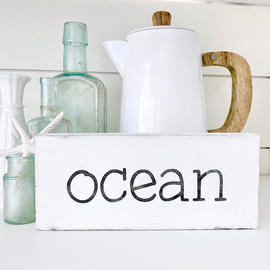 A white rectangular wooden sign with the word 'ocean' in black lowercase letters, styled on a white surface with a white enamel coffee pot, vintage glass bottles, and a starfish.