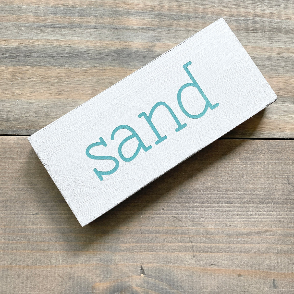 A rectangular white wooden sign with the word 'sand' painted in a light aqua lowercase font, shown on a rustic brown wood plank surface.