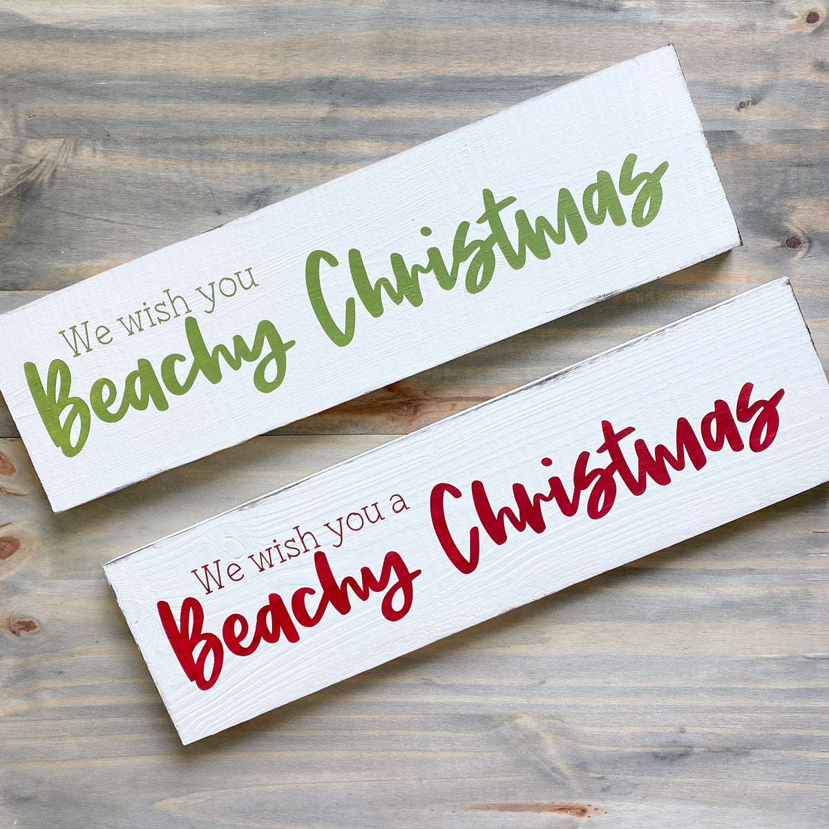 Two white rectangular wooden signs with Christmas text, displayed diagonally on a rustic gray wood background. Both read 'We wish you Beachy Christmas' in green and red lettering.