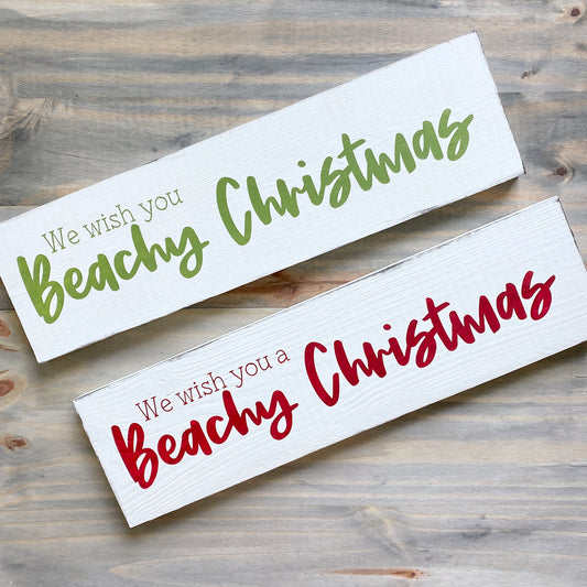 Two white rectangular wooden signs with Christmas text, displayed diagonally on a rustic gray wood background. Both read 'We wish you Beachy Christmas' in green and red lettering.