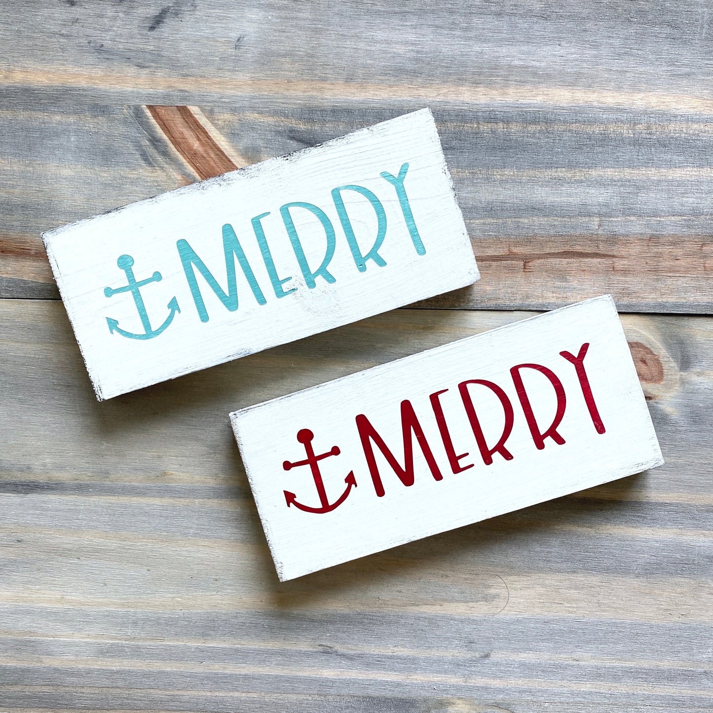Two rectangular white wooden signs with a distressed finish, one painted aqua blue and one painted red, each featuring the word “MERRY” with an anchor symbol. The signs are arranged diagonally on a rustic gray wood plank surface.