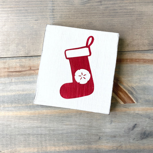 A small white square sign or plaque featuring a red Christmas stocking with a white sand dollar detail, photographed on a rustic gray-brown wooden surface.