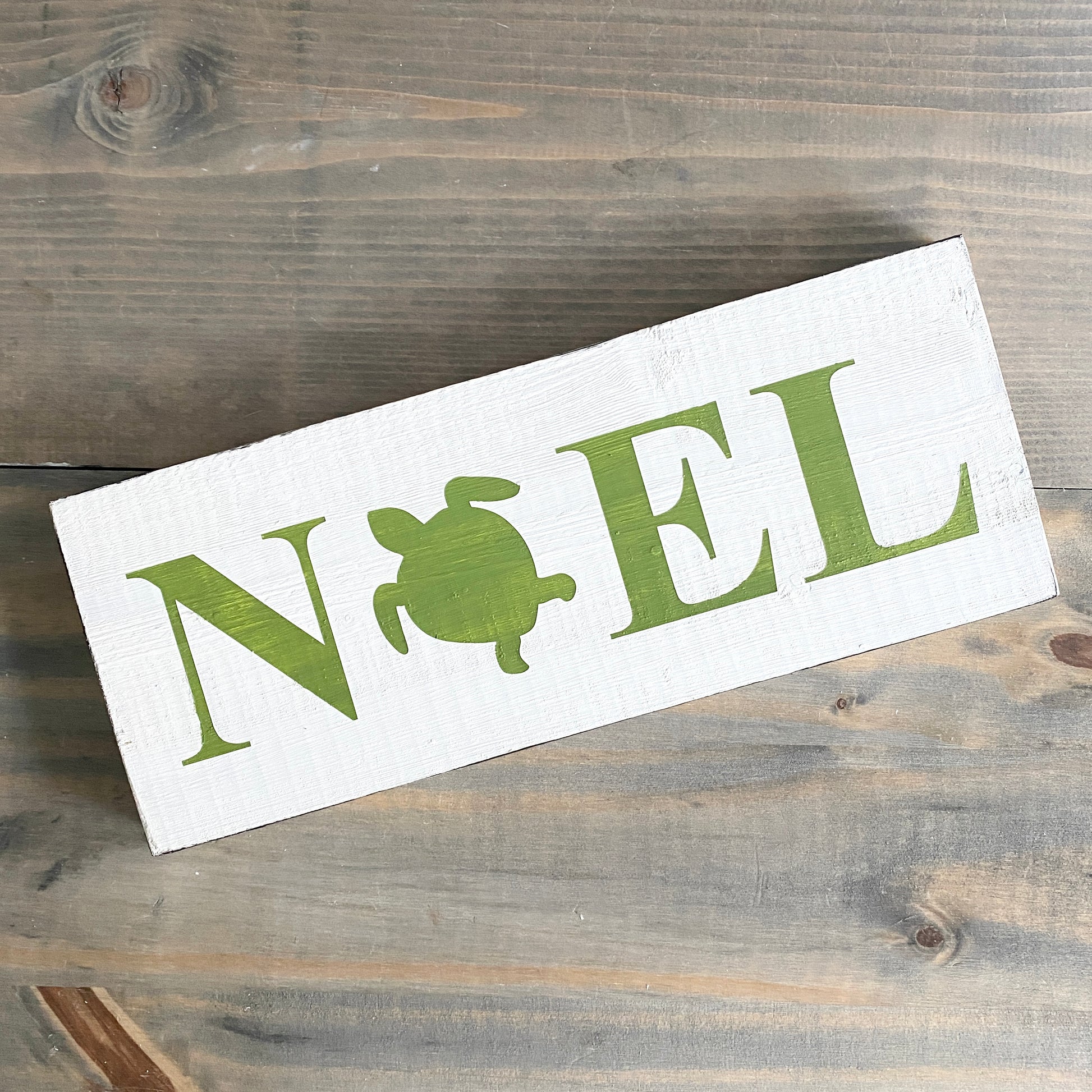 A rectangular white wooden sign with the word 'NOEL' in green, where the letter 'O' is replaced by a green turtle silhouette, displayed on a rustic gray-brown wooden surface.