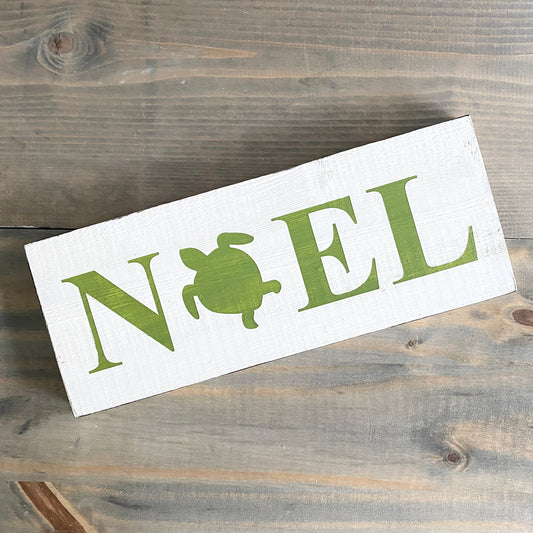 A rectangular white wooden sign with the word 'NOEL' in green, where the letter 'O' is replaced by a green turtle silhouette, displayed on a rustic gray-brown wooden surface.