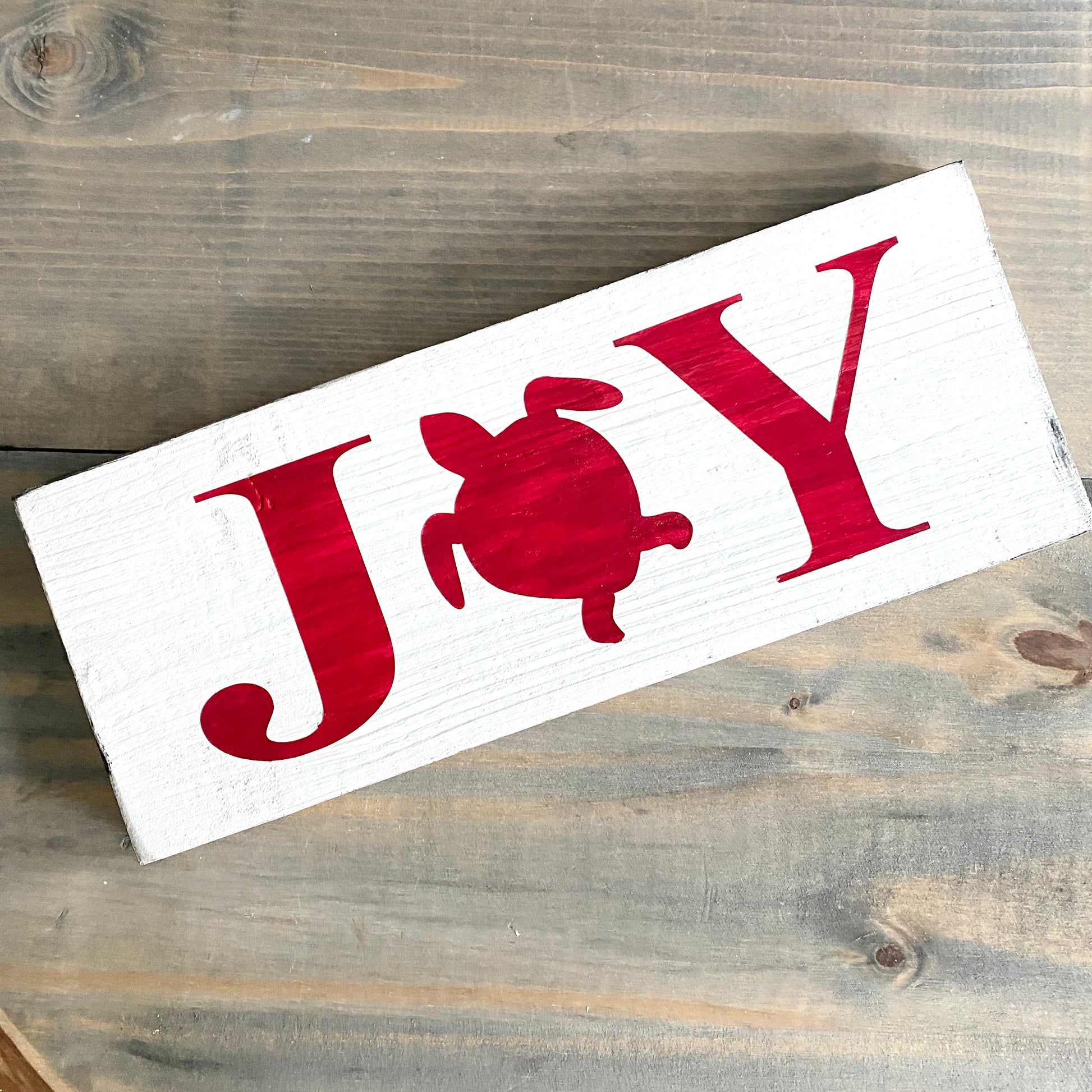 A rectangular white wooden sign with the word "JOY" in red, where the letter O is replaced by a red turtle silhouette, photographed on a rustic gray-brown wood surface.