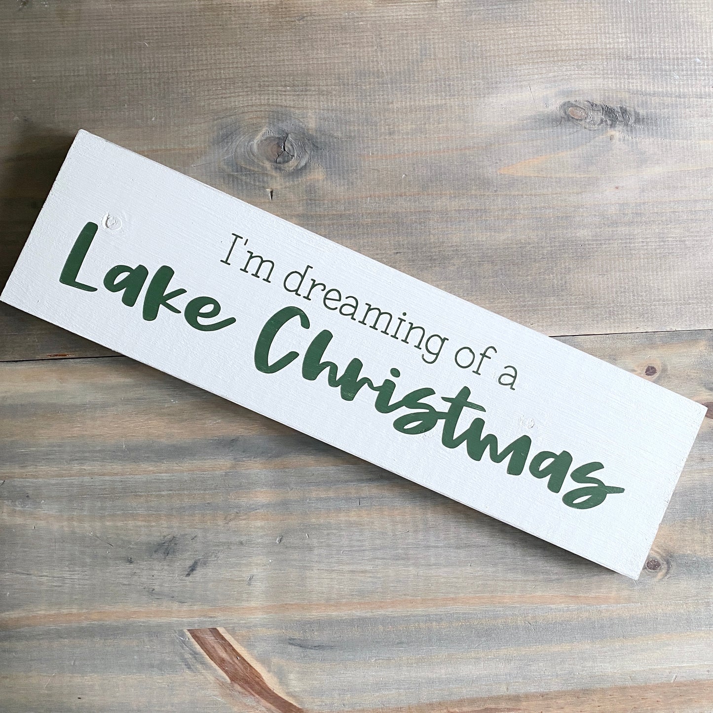A white rectangular wooden sign with the phrase 'I'm dreaming of a Lake Christmas' in dark green lettering, photographed on a rustic gray-brown wood plank surface.