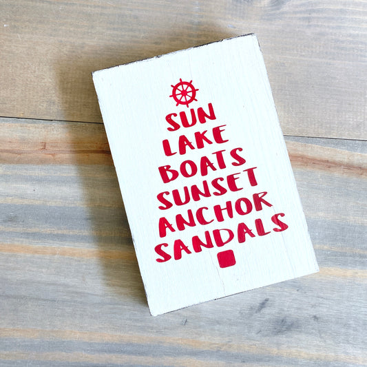 A white rectangular wooden sign with red text reading 'SUN LAKE BOATS SUNSET ANCHOR SANDALS' and a small red nautical wheel icon at the top, photographed on a gray-brown wood plank surface.