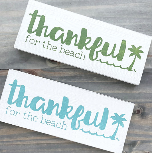 two white signs on a wood background with text- thankful for the each and a palm tree and wave, one in green, one in aqua