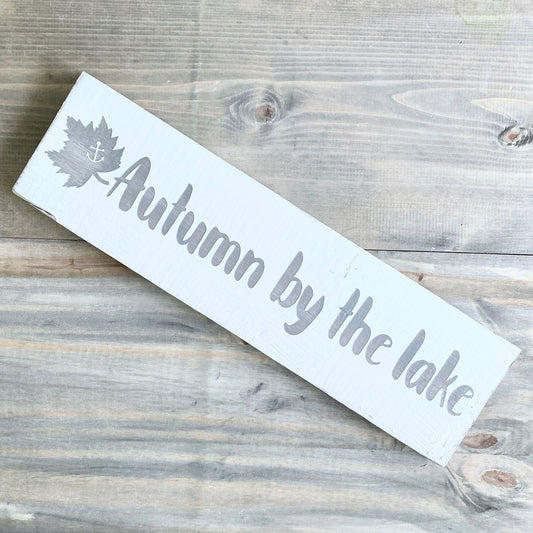 White sign with 'Autumn by the lake' text and leaf design on a wooden surface