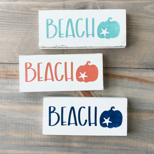 Three 'BEACH' signs with pumpkins and starfish on a wooden surface