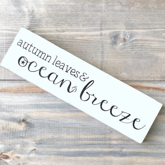 White wooden sign with 'autumn leaves & ocean breeze' text on a wooden surface