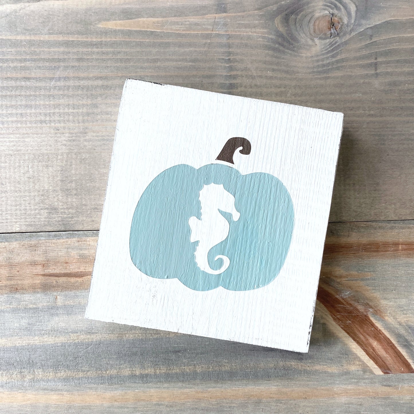 A square white wooden sign featuring a painted sage green pumpkin with a white seahorse silhouette in the center and a dark brown stem, displayed on a rustic wood plank surface.