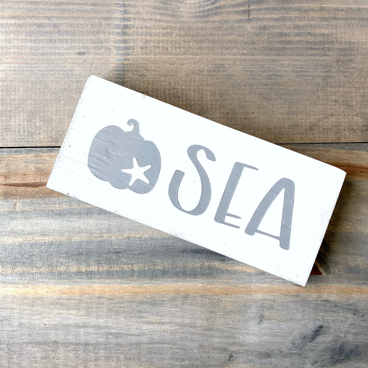 A rectangular white wooden sign with a gray painted pumpkin with starfish and the word 'SEA' in large gray letters, placed diagonally on a rustic wood plank surface.