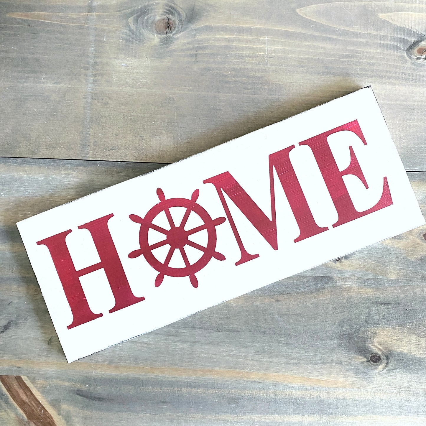 A rectangular white wooden sign with the word "HOME" in red, where the letter O is replaced by a red ship's wheel, photographed on a rustic gray-brown wood plank surface.