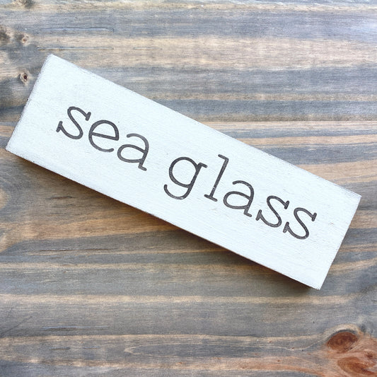 A rectangular white wooden sign with the words 'sea glass' in a black font, resting diagonally on a rustic brown wood plank surface.