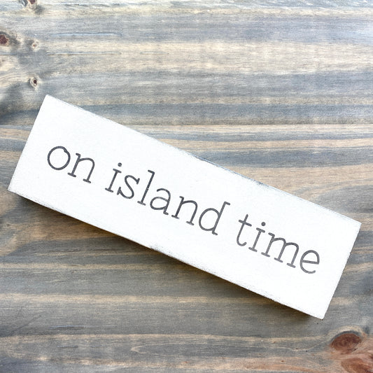 A rectangular white wooden sign with the phrase 'on island time' in black lowercase serif lettering, photographed on a rustic weathered wood surface.