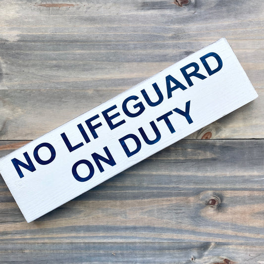A rectangular white wooden sign with the text 'NO LIFEGUARD ON DUTY' in bold navy blue lettering, shown diagonally on a rustic gray wood plank background.